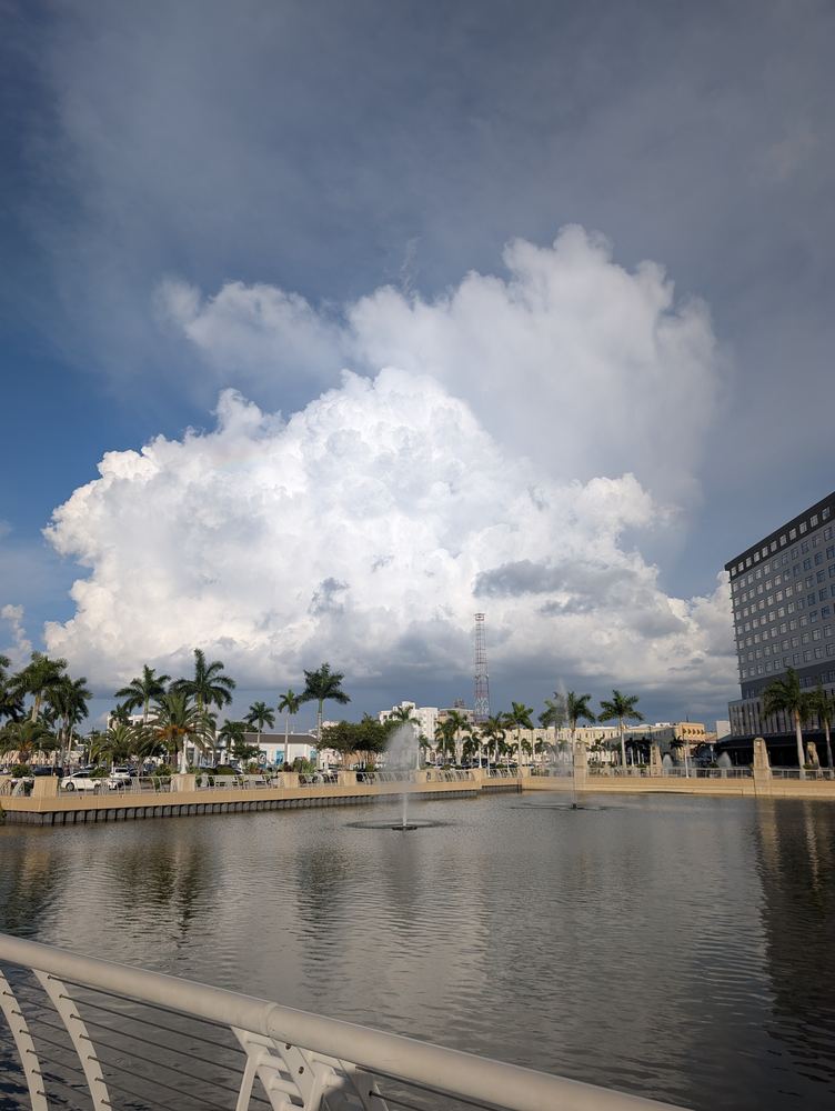 A large cumulus cloud building over downtown Fort Myers, with palm trees and a fountain pond in the foreground