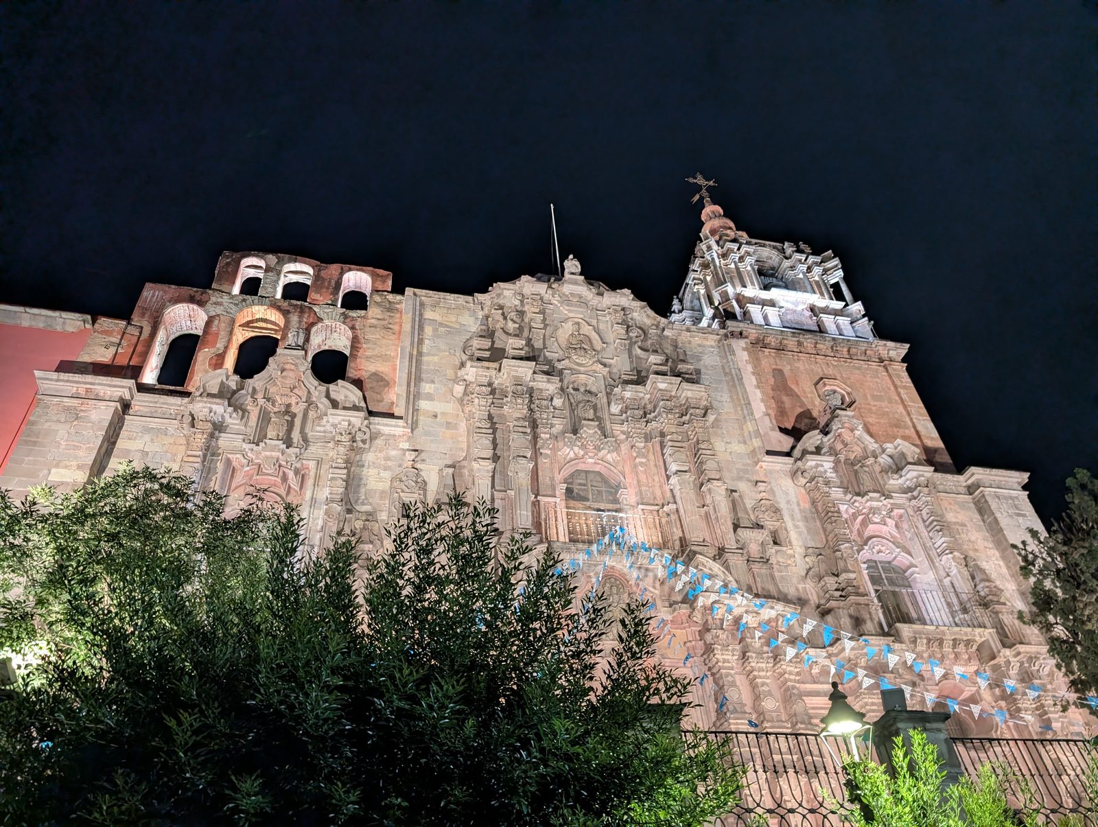 A weathered pink-stone baroque church facade strung with banners, lit up at night in Guanajuato