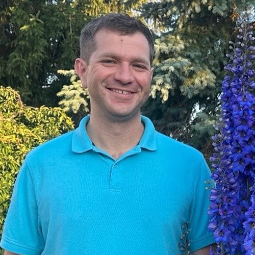 Karl Kunze smiling outdoors next to a tall blue delphinium
